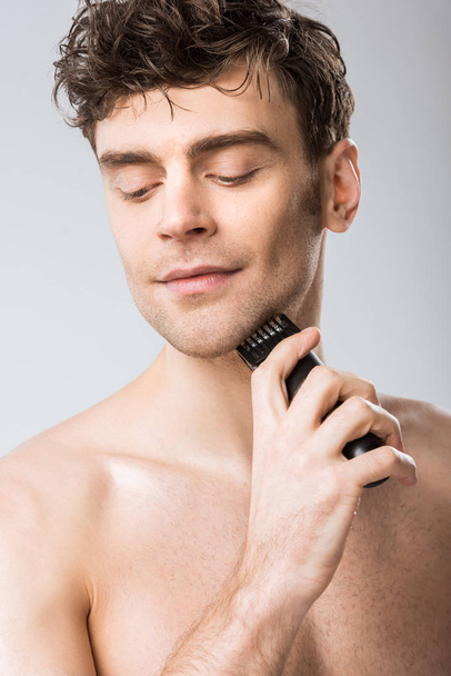 young man shaving with electric razor isolated on grey - Φωτογραφία, εικόνα