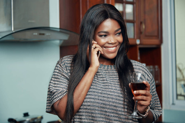 Весела чорна жінка тримає келих вина і розслабляється вдома, розмовляючи на смартфоні
 - Фото, зображення