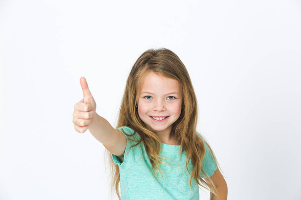 portrait of pretty little girl in green t-shirt showing thumb up while posing in studio  - Photo, image