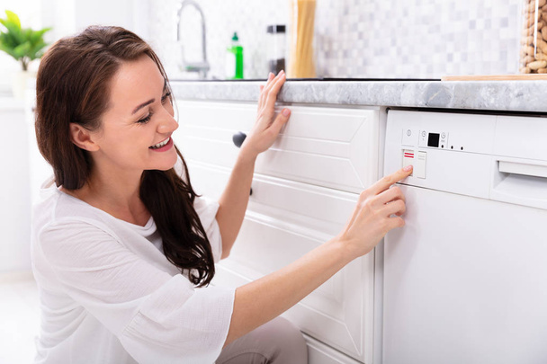 Вид сбоку на счастливую молодую женщину, нажимающую кнопку посудомоечной машины
 - Фото, изображение