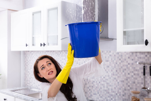 Unhappy Young Woman Collecting Water Leaking From Ceiling In Blue Bucket - 写真・画像