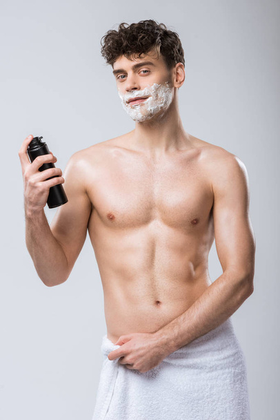 handsome shirtless young man holding bottle with shaving foam, isolated on grey - Photo, Image