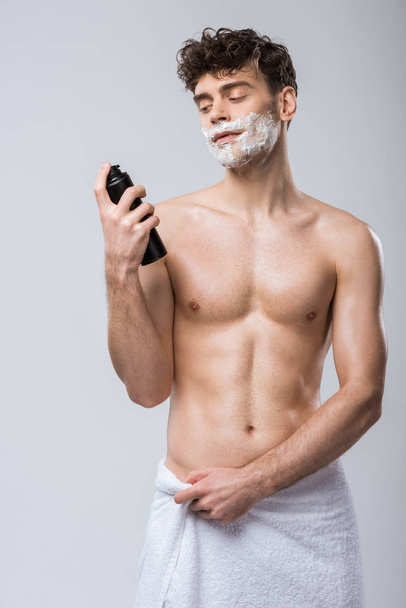 Красивый молодой человек в полотенце держит бутылку с пеной для бритья, изолированный на сером
 - Фото, изображение