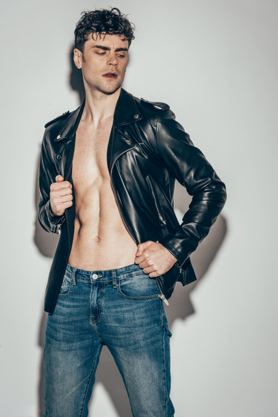 handsome sexy rocker posing in jeans and black leather jacket on grey - Foto, afbeelding