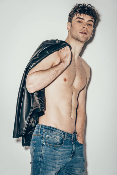 σέξι άνδρας shirtless ποζάρει με το μαύρο δερμάτινο μπουφάν σε γκρι - Φωτογραφία, εικόνα