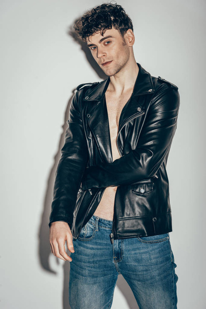 stylish sexy man posing in jeans and black leather jacket on grey - Foto, Imagen