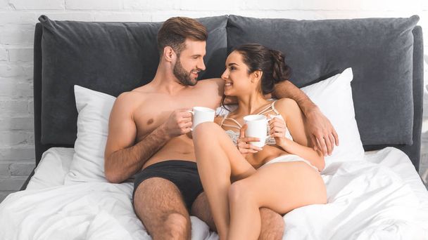 nádherný sexy pár ležící s šálky s kávou v posteli, s úsměvem a při pohledu na sebe - Fotografie, Obrázek