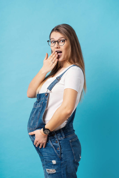 Portrait of good-looking young, cheerful pregnant woman wearing comfy denim romper and glasses making surprised face, looking at camera with a happy face expression over blue background. - Photo, Image