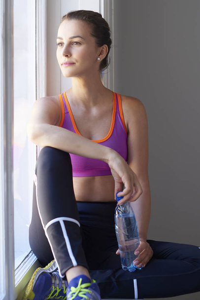 Portrait of a beautiful young woman holding a bottle of water in her hand while sitting at windown in the gym and looking out the window. - 写真・画像