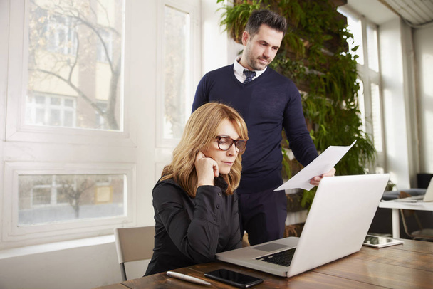 Shot of executive middle aged businesswoman using laptop while young financial assistant businessman standing next to her and working together. Teamwork in the office. - Foto, imagen