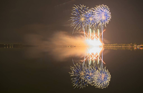 Kolorowy fajerwerk na nocnym niebie z połyskujące wody refleksji nad jeziorem. - Zdjęcie, obraz