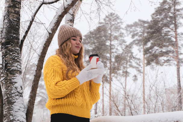 Девушка в свитере держит бумажный стаканчик с горячим напитком в зимнем пар
 - Фото, изображение