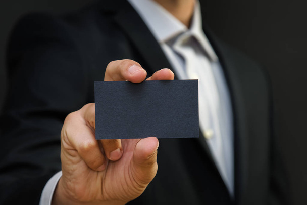 Man wearing a suit holding white business card on black wall background. Mock up, space for text - Foto, Imagem