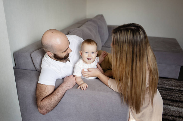 Smiling mother and father holding their baby girl daughter sitting on bed at home. Happy family concept, Bearded man in white shirt, cheerful woman hugging a child - Photo, Image