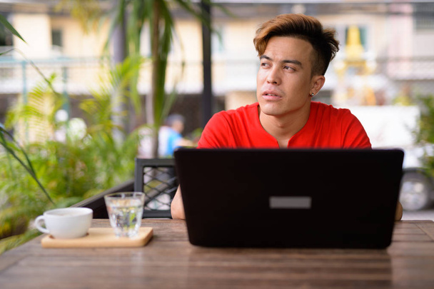 Young Asian man thinking while using laptop at the coffee shop outdoors - Photo, Image