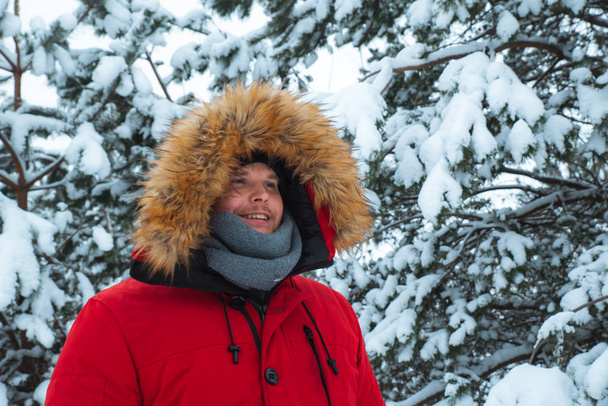 Πορτραίτο νεαρού ενήλικα σε κόκκινο χειμερινό παλτό με κουκούλα με γούνα. σε φόντο χιονισμένου έλατο. - Φωτογραφία, εικόνα