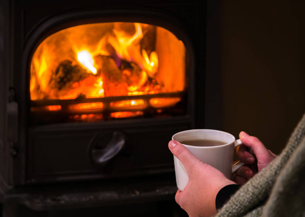 Woman having a hot cup of rink at the fireplace. Burning fireplace with wooden logs burning inside in the background. Warm light, romantic, christmas like atmosphere  - Photo, Image
