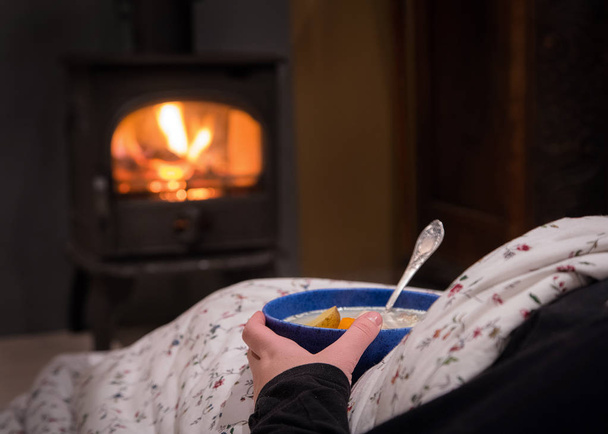 Γυναίκα έχοντας ένα ζεστό μπολ σούπα λαχανικών στο τζάκι. Καίγοντας εστία με τα ξύλινα κούτσουρα που καίει μέσα στο παρασκήνιο. Θερμό φως, Ρομαντικό, Χριστουγεννιάτικη ατμόσφαιρα  - Φωτογραφία, εικόνα