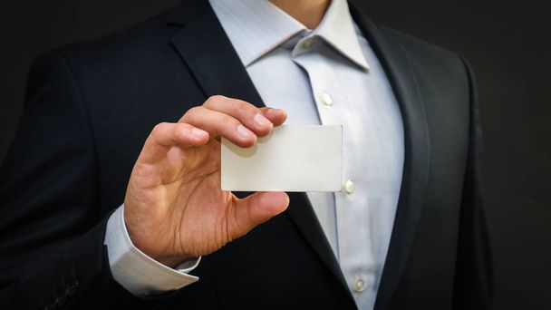 Man wearing a suit holding white business card on black wall background. Mock up, space for text - Фото, зображення