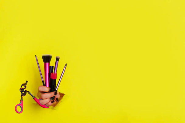 Woman hand with professional cosmetic tools for make up: brushes, mascara, lipstick, eyelash curler on yellow background. Beauty concept. Banner for cosmetics sale. Copy space. - Foto, imagen