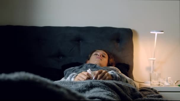 Mujer joven enferma con fiebre estornudando por la noche en la cama. Medicamentos, termómetro y vaso de agua sobre la mesa
. - Imágenes, Vídeo