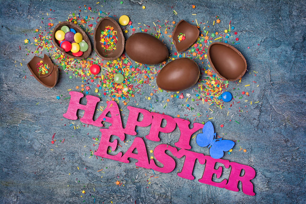 Top view on pink text of happy easter and chocolate traditional eggs with bright colorful dragee and sugar sprinkles or confetti on blue concrete background - Photo, image