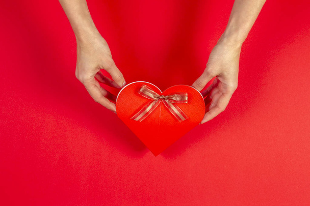 Femate hands holding gift box in heart shape with bow on red background - Zdjęcie, obraz