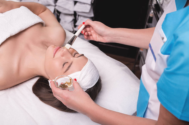 Молодая женщина наслаждается процедурой для лица в салоне красоты. Девушка лежит в спа и получает глиняную маску с удовольствием
 - Фото, изображение