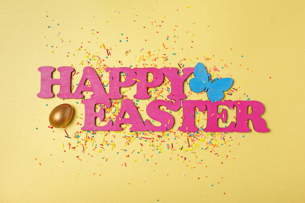 Top view on pink text of happy easter and golden painted egg with bright colorful sugar sprinkles or confetti as baking decor on yellow background - Photo, image