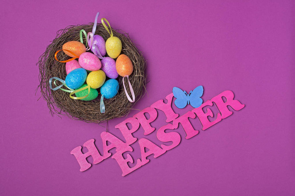 Bovenaanzicht op roze tekst van vrolijk Pasen en heldere kleurrijke glanzende eieren in het nest van de vogel op paarse achtergrond - Foto, afbeelding