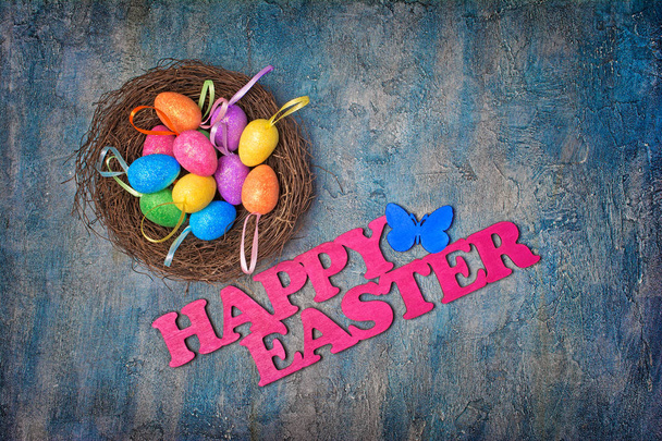 Top view on pink text of happy easter and bright colorful shiny eggs in bird nest on blue concrete background - Photo, Image