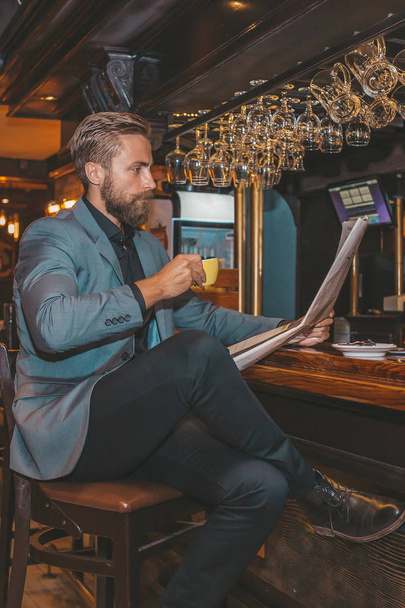 バーに座って、新聞を読んだり、コーヒーを飲む若い髭があるビジネスマンの肖像画.  - 写真・画像