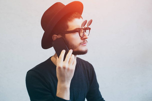 Портрет серйозного молодого хлопця, який використовує смартфон на білому тлі. Сонцезахисні окуляри та чорний капелюх
 - Фото, зображення