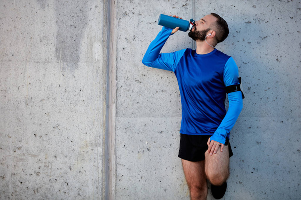 Man resting from running and drinking water while standing against the wall. Healthy lifestyle concept. - Фото, изображение