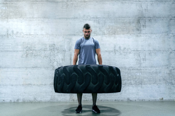 starker muskulöser Mann in Sportbekleidung, der Reifen hebt, während er in der Crossfit-Turnhalle vor der Wand steht. - Foto, Bild