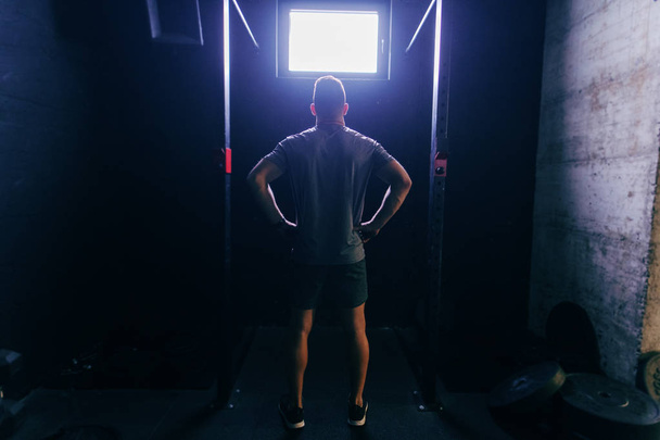 muskulöser Mann in Sportbekleidung, der sich auf Klimmzüge vorbereitet, während er mit den Händen auf den Hüften im Crossfit-Fitnessstudio steht. Rückwärts gedreht. - Foto, Bild