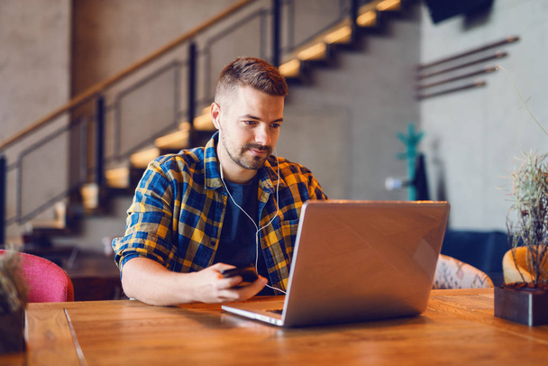 Freelancer jovem caucasiano com sorriso no rosto e fones de ouvido nos ouvidos trabalhando no laptop enquanto sentado na cafetaria
. - Foto, Imagem