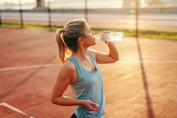 sportliche kaukasische Blondine steht auf dem Platz und trinkt Wasser nach morgendlichen Übungen. - Foto, Bild
