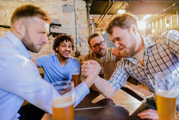 jóvenes hombres barbudos felices bebiendo con cerveza en el pub y teniendo desafío de lucha libre brazo
  - Foto, Imagen