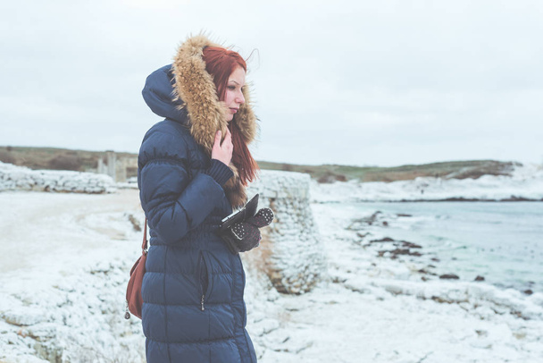 Girl in a warm jacket with fur stands on the frozen bank of the winter sea. - Fotografie, Obrázek