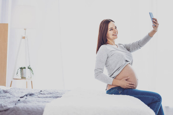 Happy pregnant woman taking selfie while sitting on bed - Zdjęcie, obraz