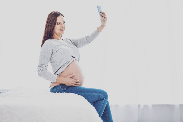 Cheerful pregnant woman posing for selfie on bed - Фото, изображение