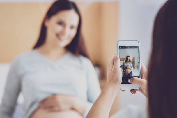 Preteen girl taking photo of pregnant woman sitting on bed - Foto, Imagen