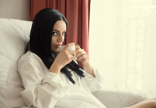 Girl with black hair is sitting next to the window and drinking tea - Foto, imagen