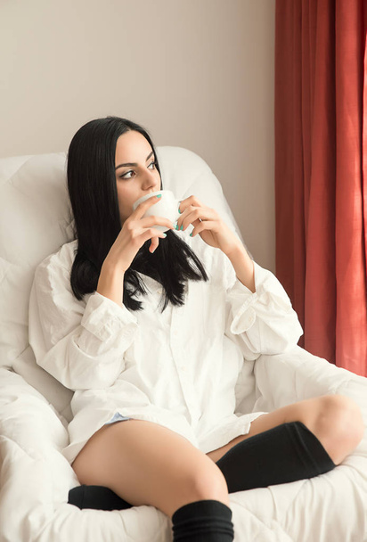 Girl with black hair is sitting next to the window and drinking tea - Zdjęcie, obraz