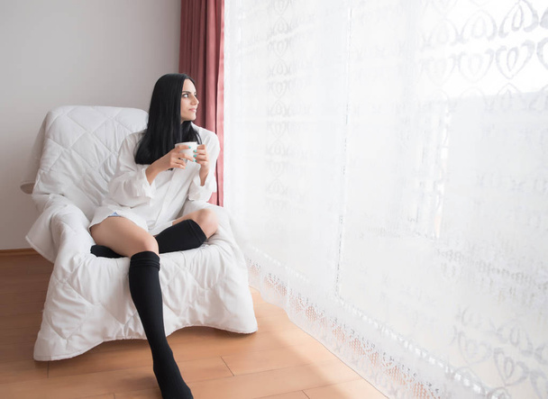 Girl with black hair is sitting next to the window and drinking tea - Φωτογραφία, εικόνα