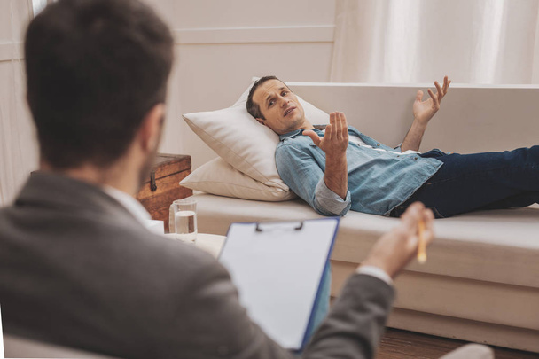 Gestresster Mann liegt auf beigem Sofa und erklärt Therapeut seine Probleme - Foto, Bild
