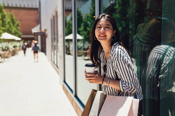 Urocza dziewczyna na atrakcyjne, uśmiechający się wyglądający stojący aparat twarz opierając się przeciwko okna poza tkaniny sklep w centrum handlowego stanford przytrzymanie relaksujący pod słońcem kawa. Azjatyckie kobiety noszenia torby. - Zdjęcie, obraz