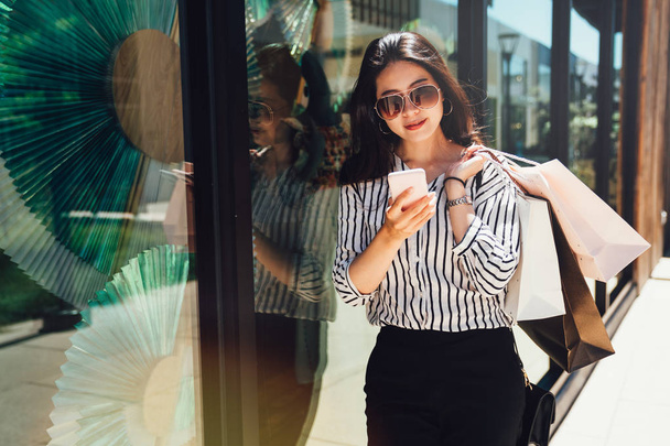 Beautiful fashionable young asian woman carrying shopping bags relying against display window of cloth shop in outdoor mall center in stanford. girl in sunglasses holding smartphone touching screen. - Photo, image
