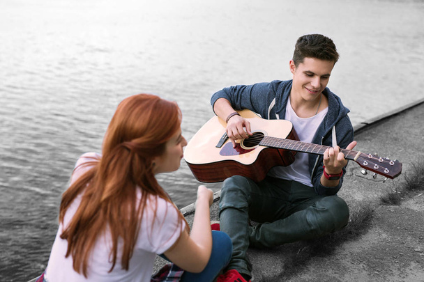 Handsome romantic boyfriend playing the guitar for his girlfriend sitting near river - 写真・画像